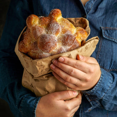 Person holding a brown paper bag with a large day of the dead bread gun wrapped in it.