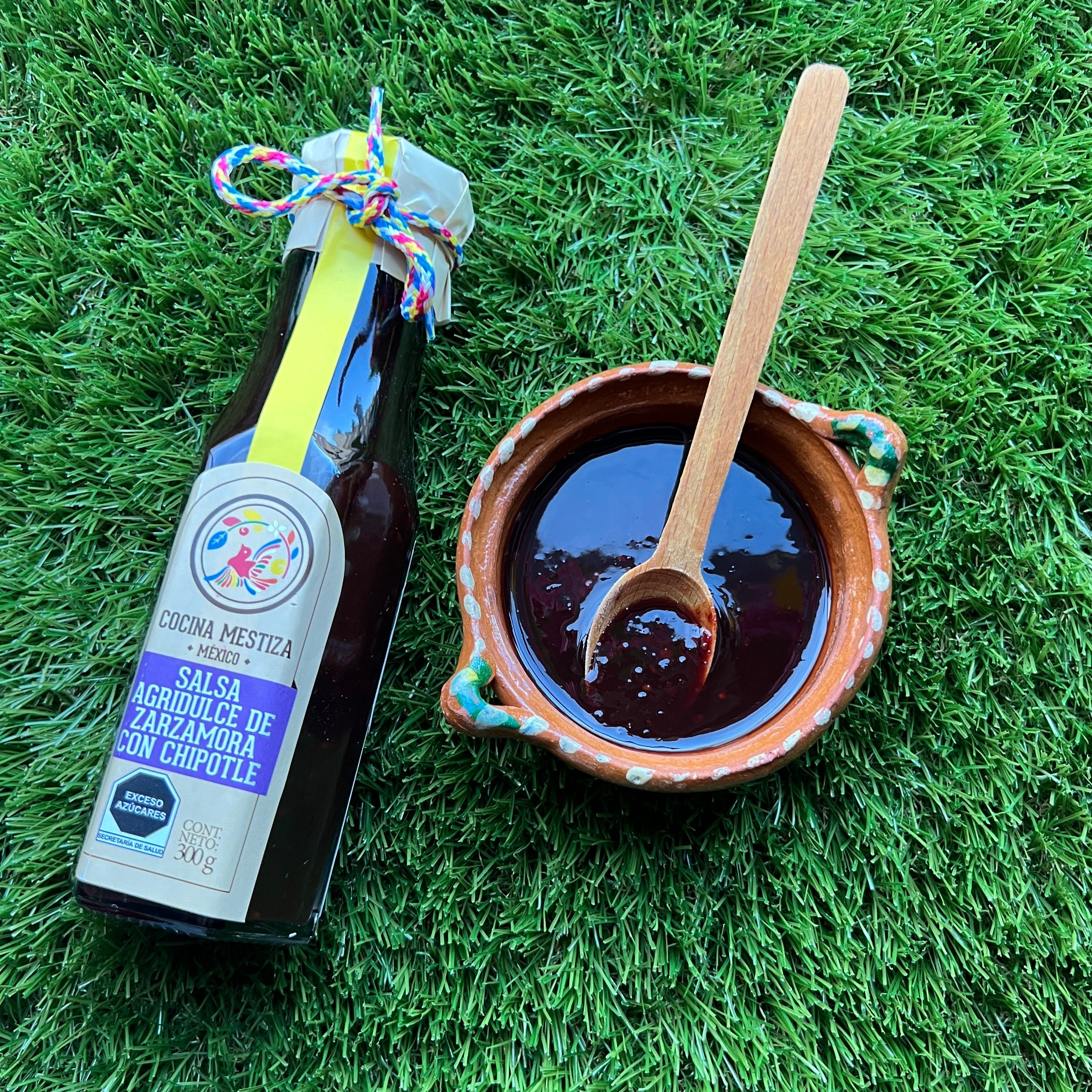 a bottle of Blackberry & Chipotle Salsa laying on the grass with a cute Mexican clay bowl with some of the salsa in it and a wooden spoon