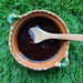 A small Mexican Clay bowl filled with some Blackberry & Chipotle Salsa laying on the grass. The bowl has a small wooden spoon